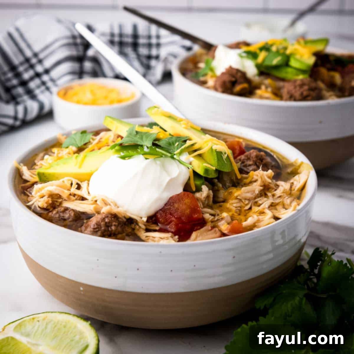 Side angled shot of two white bowls of chicken taco soup with slices of avocado, fresh cilantro, shredded cheese, and sour cream on top with lime wedges on the side.