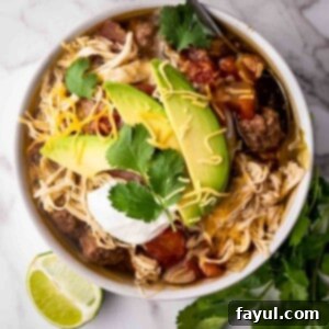 Overhead shot of chicken taco soup with slices of avocado, fresh cilantro, shredded cheese, and sour cream on top all in a white bowl with a lime wedge and fresh cilantro on the side.