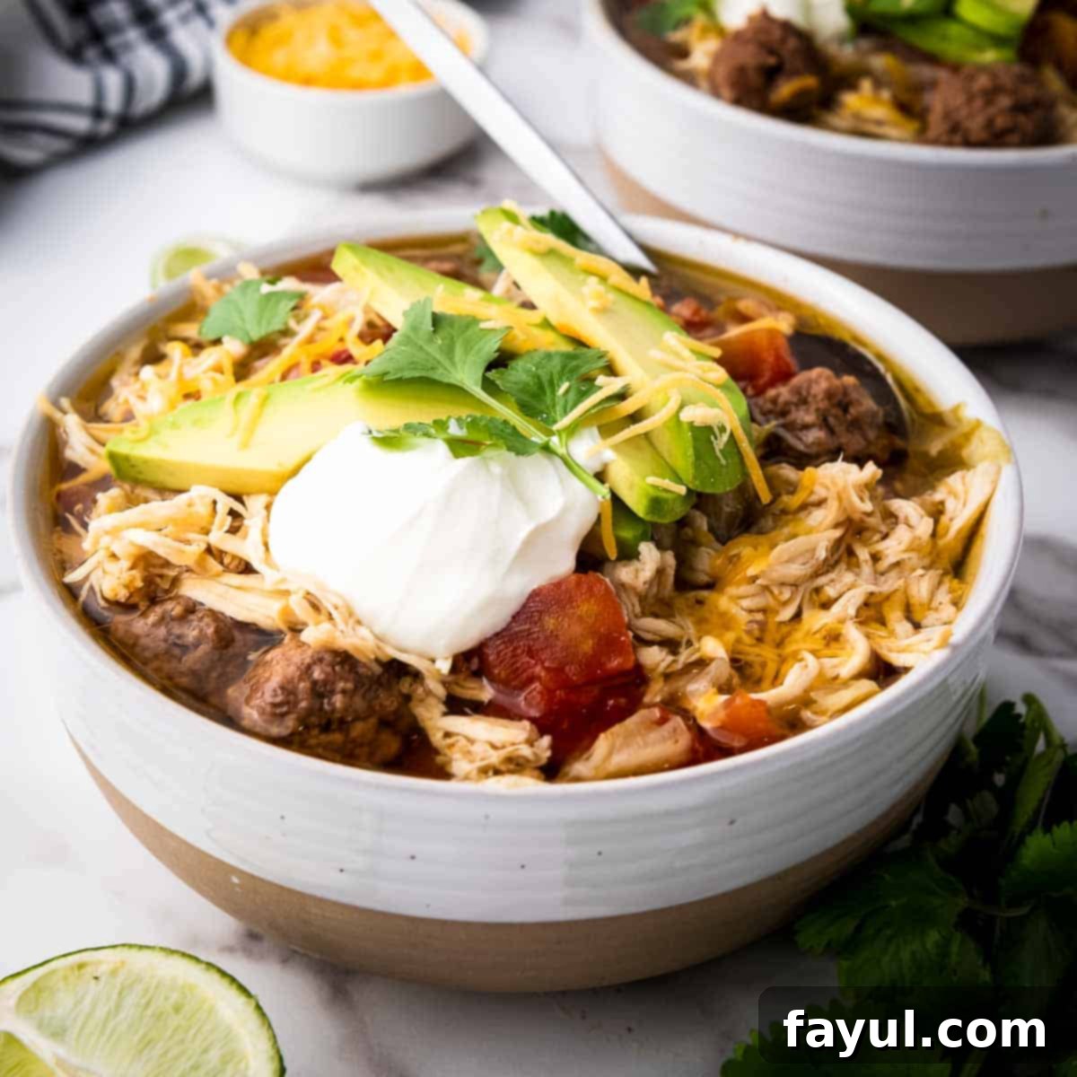 Side angled shot of chicken taco soup with slices of avocado, fresh cilantro, shredded cheese, and sour cream on top all in a white bowl.