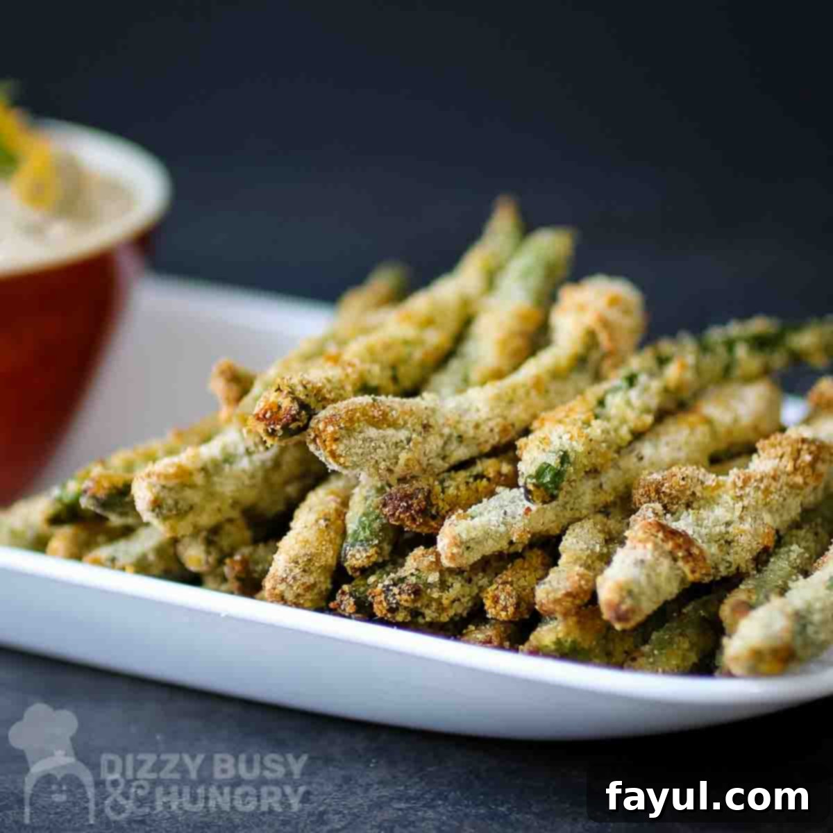 Side view of a stack of golden-brown crispy baked green bean fries on a white plate, with a red dipping bowl and a black background.