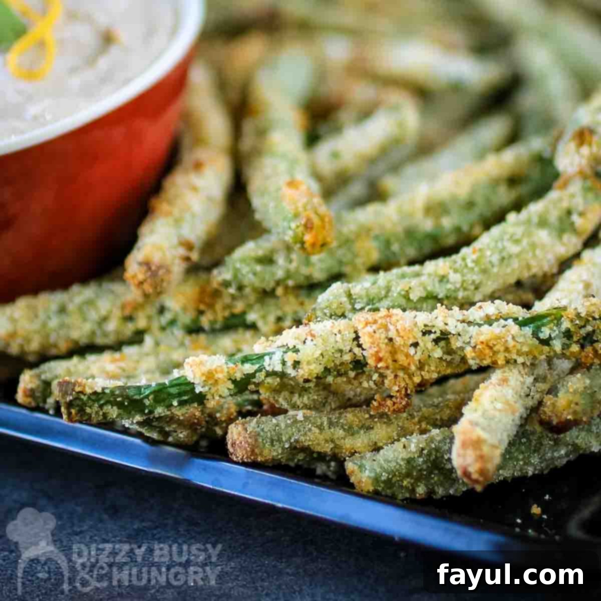Close-up side view of crispy baked green bean fries perfectly stacked on a black plate, with a red bowl in the background and a black backdrop.