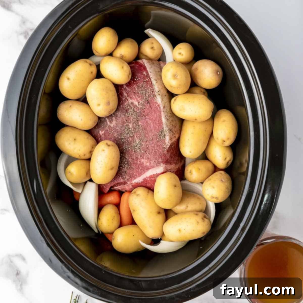 Overhead view of process shot 2: Peeled and quartered onion, baby carrots, and baby potatoes are added around the rump roast in the crock pot, creating a colorful bed of vegetables.