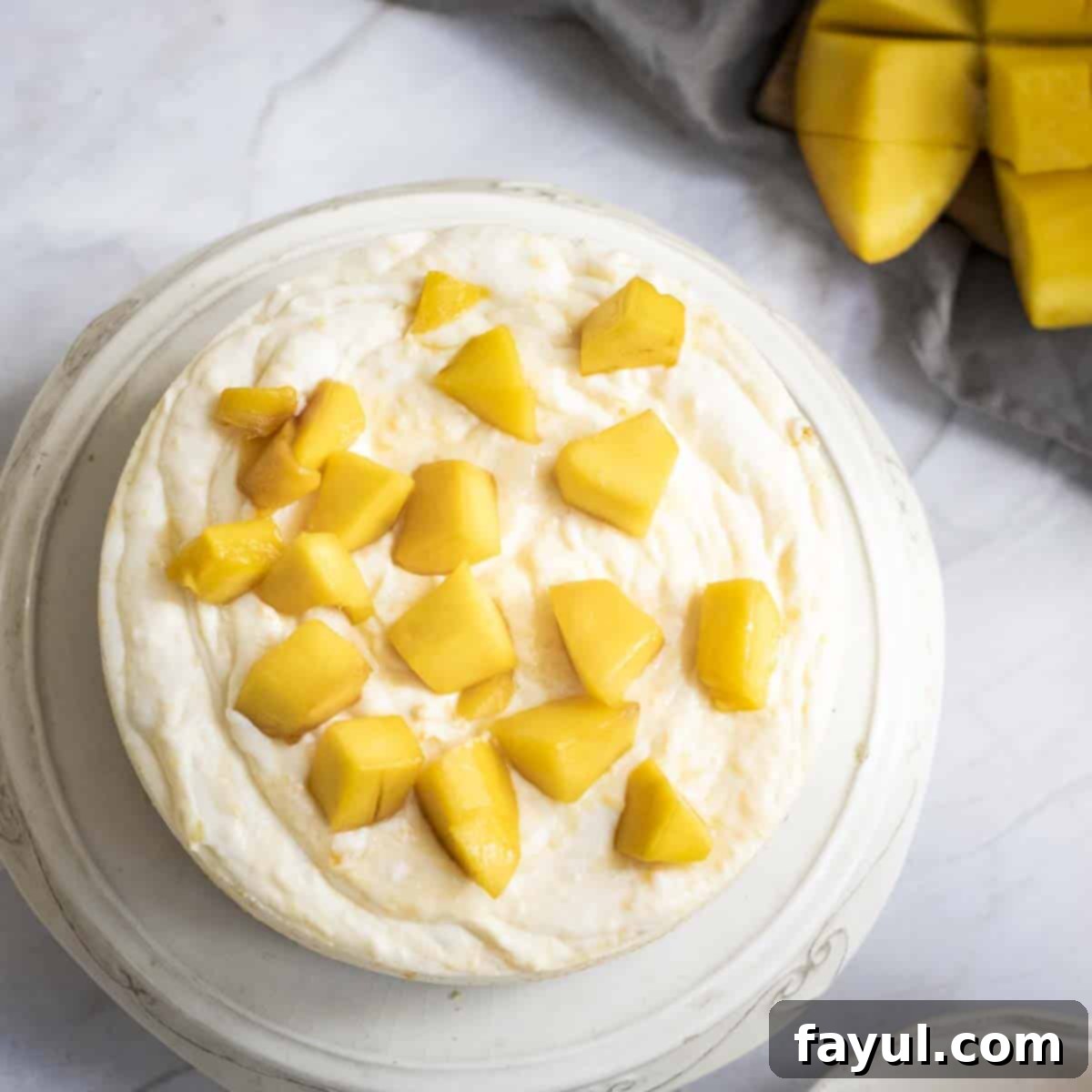 Velvet Mango No-Bake Delight 9 Overhead shot of mango cheesecake on a white decorative plate with mango chunks on top and on the side.