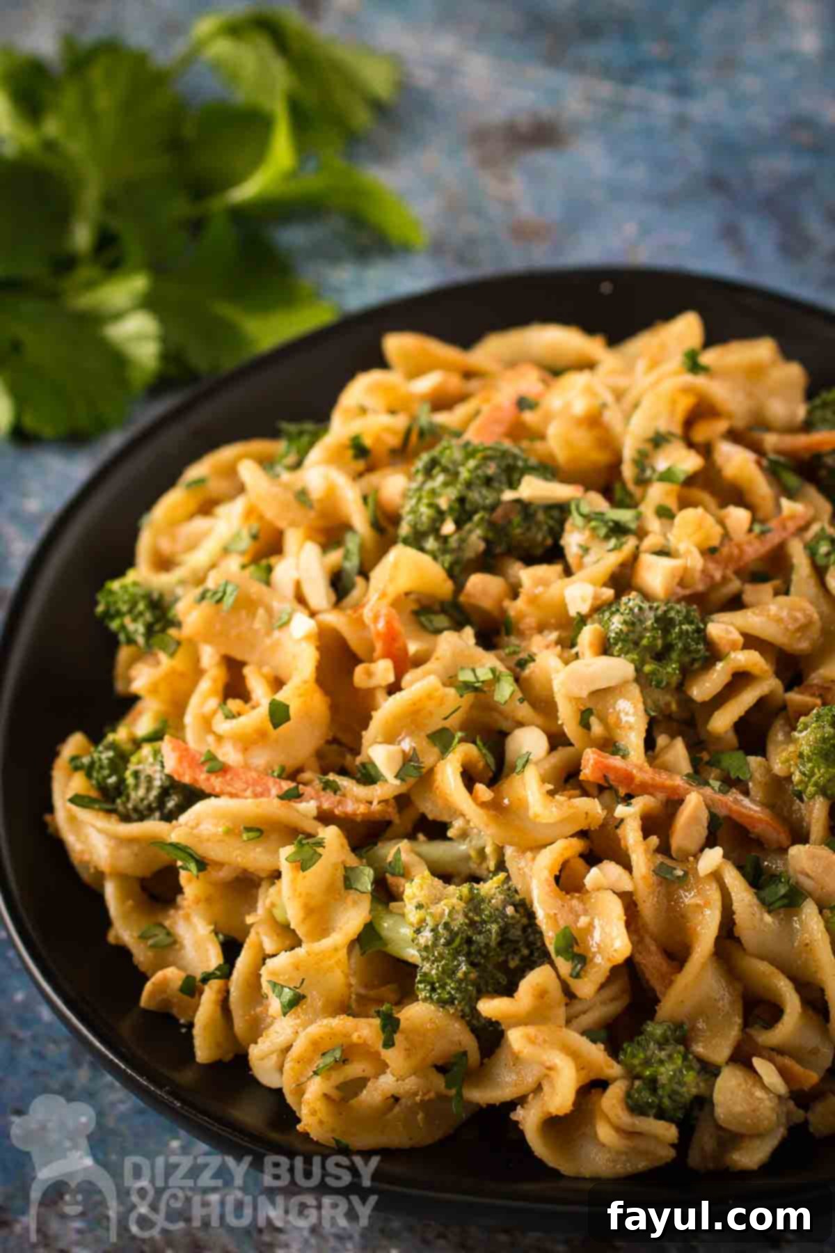 Side angled shot of noodles and broccoli garnished with chopped peanuts on a black plate with herbs on the side.
