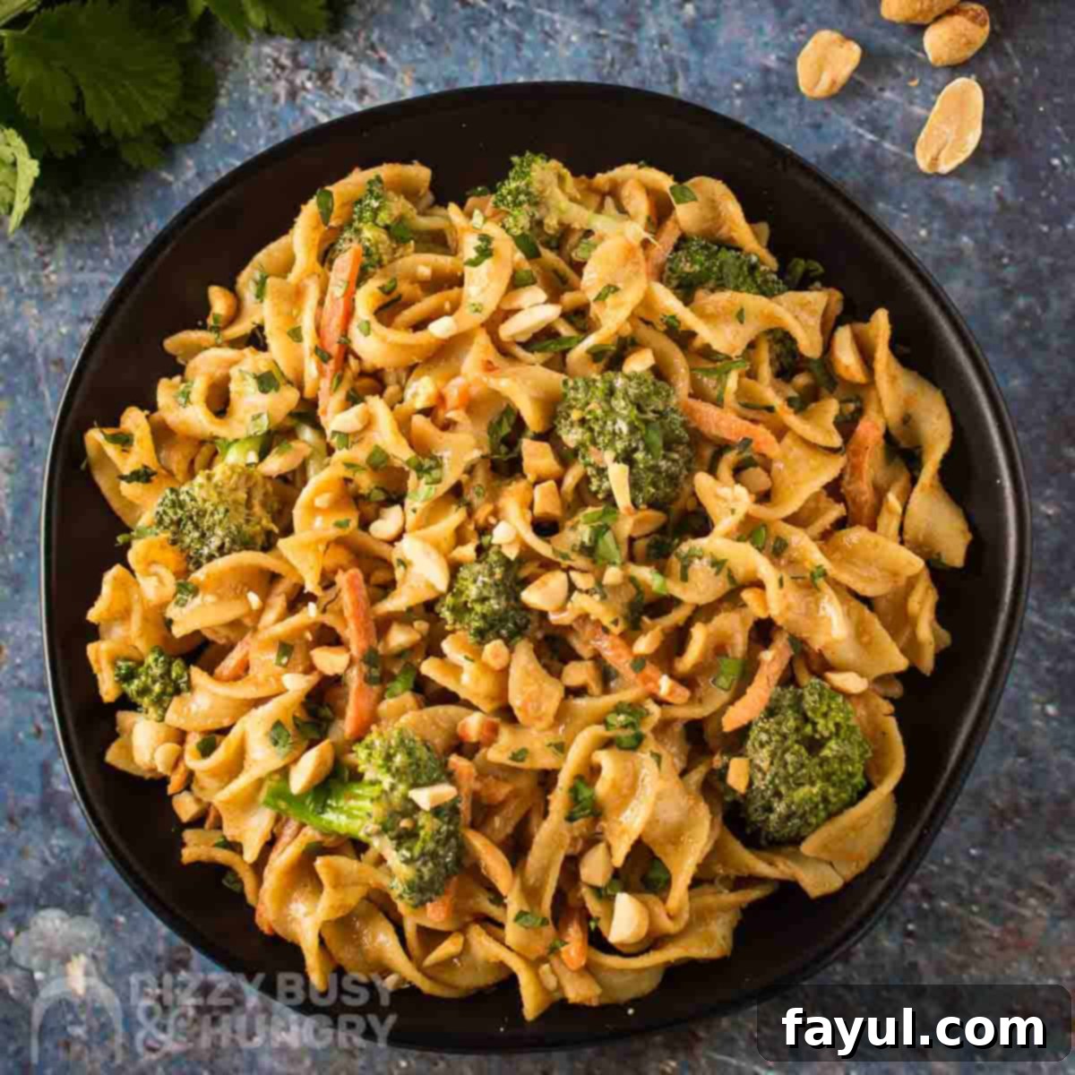 Overhead shot of noodles and broccoli garnished with chopped peanuts on a black plate with herbs and peanuts on the side.
