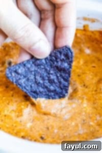 Hand dipping blue tortilla chip into chili cheese dip.