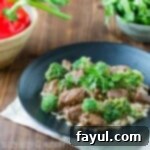 Side view of Crock Pot Beef and Broccoli on a black plate, garnished with fresh herbs and bell peppers in the background.