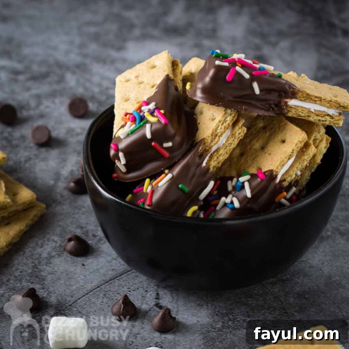 Side shot of s'mores bites in a black bowl on a grey surface with marshmallows, chocolate chips, and graham crackers on the side.