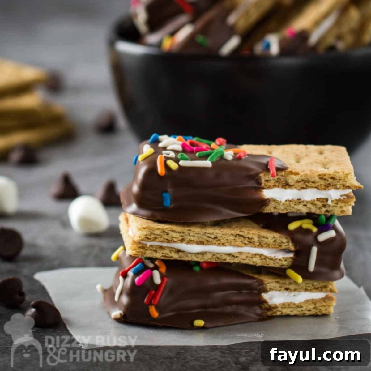 Side shot of three s'mores bites stacked on each other on a grey surface with a black bowl in the background and marshmallows and chocolate chips scattered around.