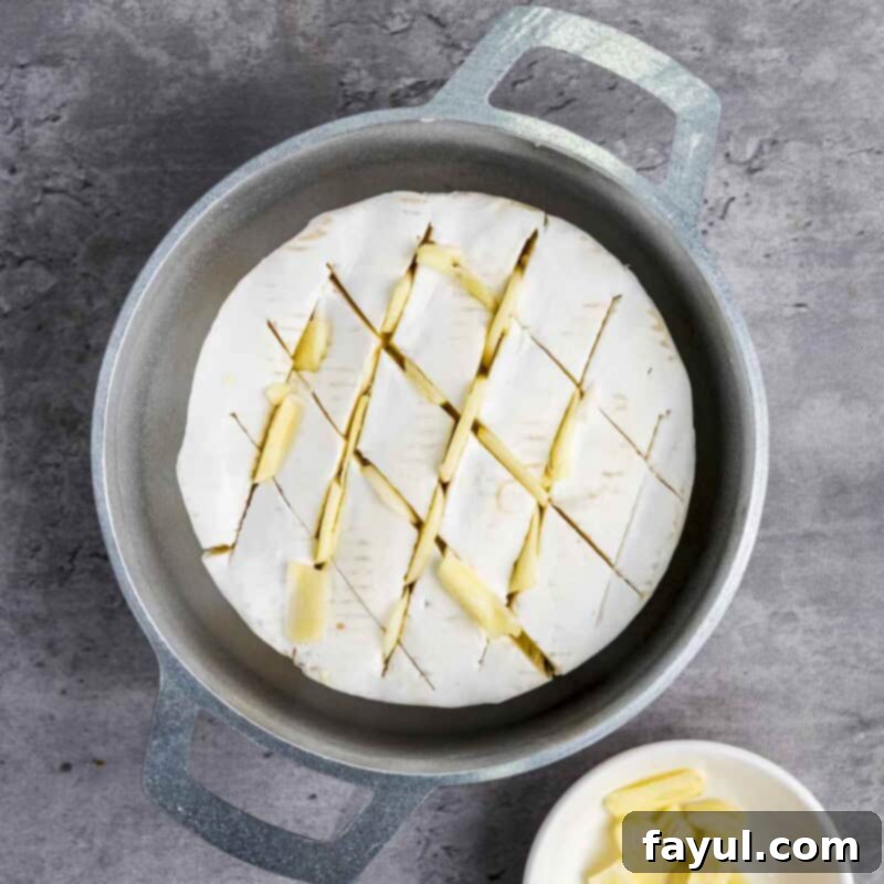 Overhead view of process shot 1: a wheel of brie cheese placed in a small baking dish, with small slits cut into its top surface. Thin slices of garlic are being inserted into these slits, distributing them evenly across the cheese for flavor infusion.