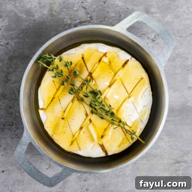 Overhead view of process shot 3: fresh thyme sprigs artfully placed on top of the honey-drizzled brie in a baking dish, just before it goes into the oven to bake until soft and gooey and aromatic.