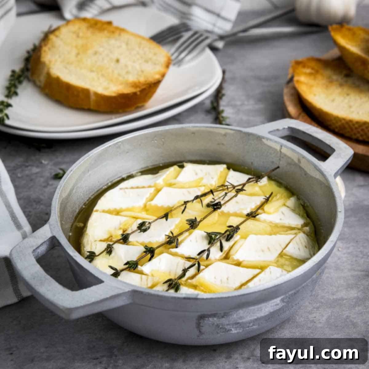 Side shot of warm baked brie garnished with fresh thyme in a small baking dish, surrounded by more thyme sprigs and toasted bread for dipping.
