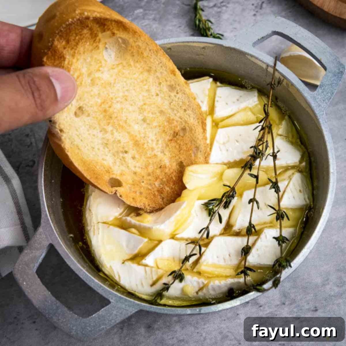 Close up shot of toasted bread being dipped into warm, gooey baked brie, garnished with fresh rosemary sprigs, all nestled in a small baking dish, highlighting the inviting texture.