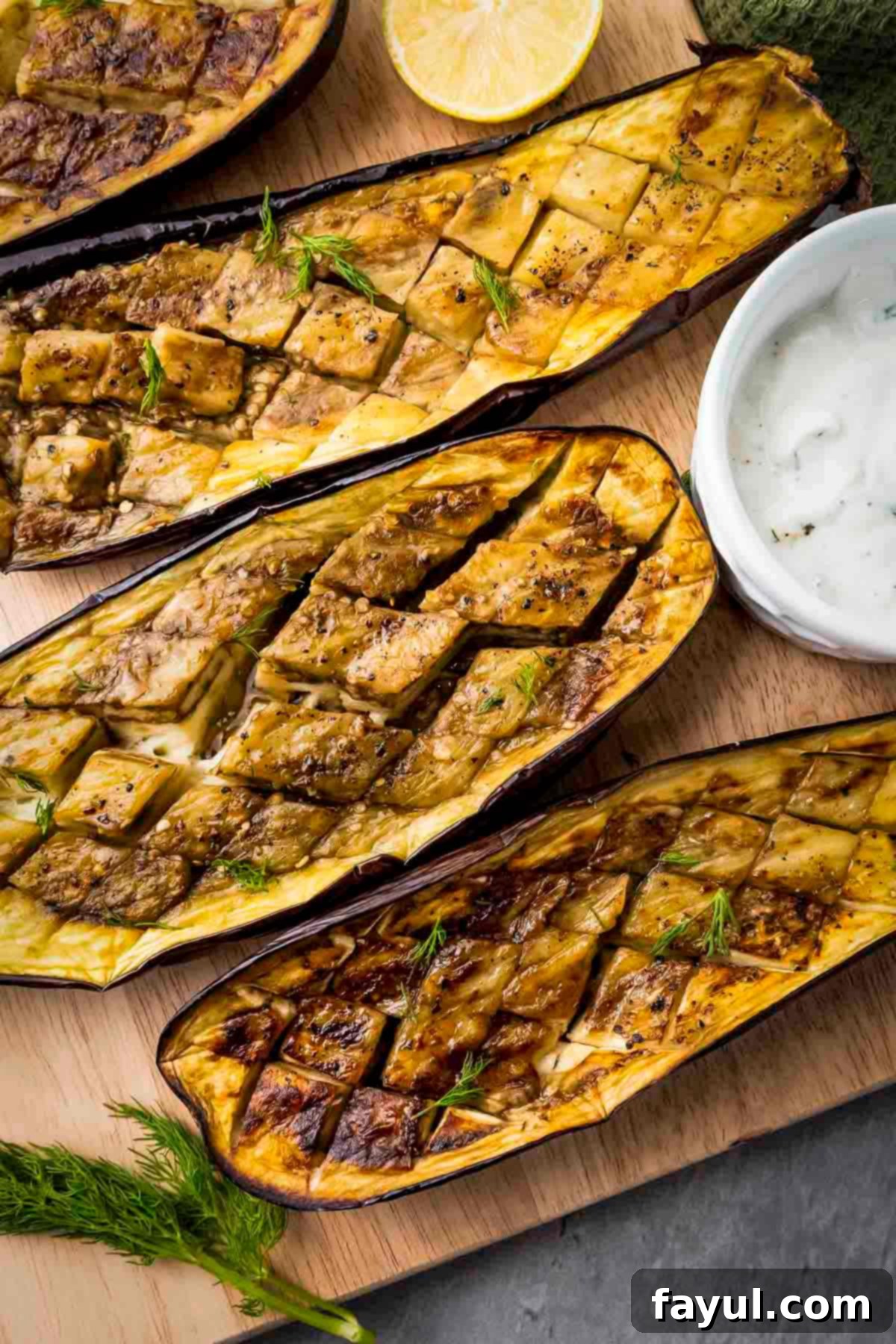Overhead shot of four slices of roasted eggplant on a wooden cutting board with fresh dill, lemon slices, and dipping sauce on the side. The eggplant is golden-brown and garnished with fresh herbs, ready to be served with the creamy white sauce.