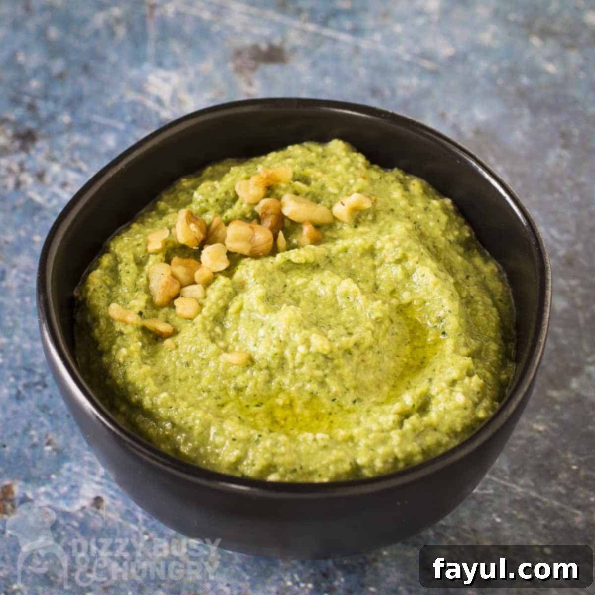 Side shot of zucchini pesto garnished with chopped walnuts in a black bowl on a blue marble surface.