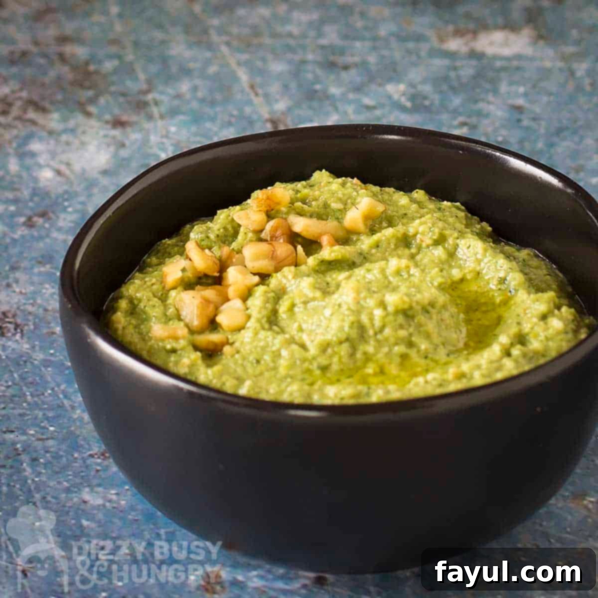 Side shot of zucchini pesto garnished with chopped walnuts in a black bowl on a blue marble surface.