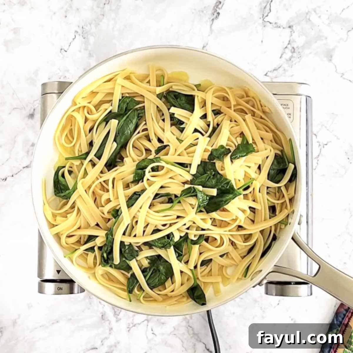 Overhead view of process shot 4 - Cooked fettuccine and wilted spinach being added to the skillet with the garlic and olive oil, then tossed to combine thoroughly.