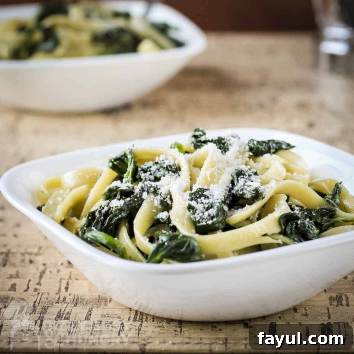Side close up shot of a fork twirling fettuccine with spinach and parmesan in a white bowl on a wooden table, showcasing the texture and vibrant greens.
