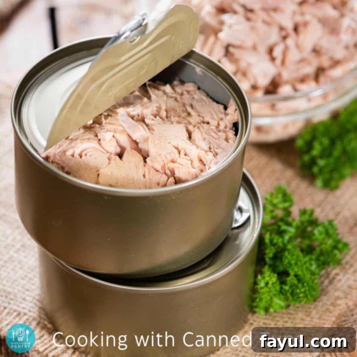 Partially opened can of tuna on top of another can of tuna with parsley on the right side.
