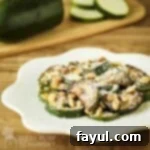 Side shot of sauteed zucchini and walnuts with parmesan cheese on a white decorative plate with raw zucchini on a cutting board in the background.