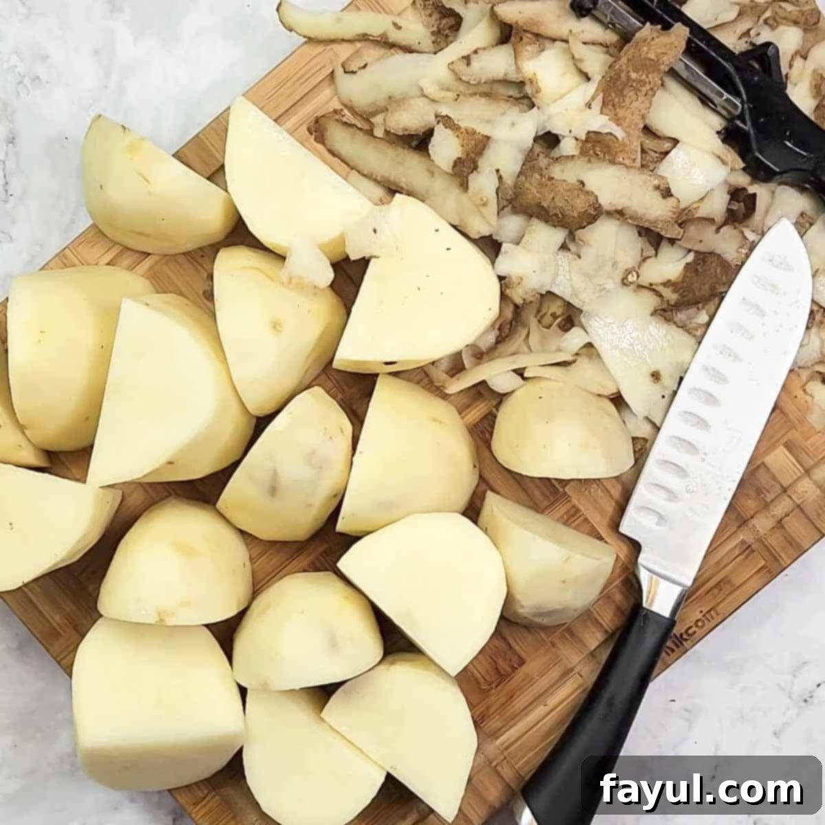 Overhead view of potatoes cut into chunks on a wooden cutting board next to a cutting knife.