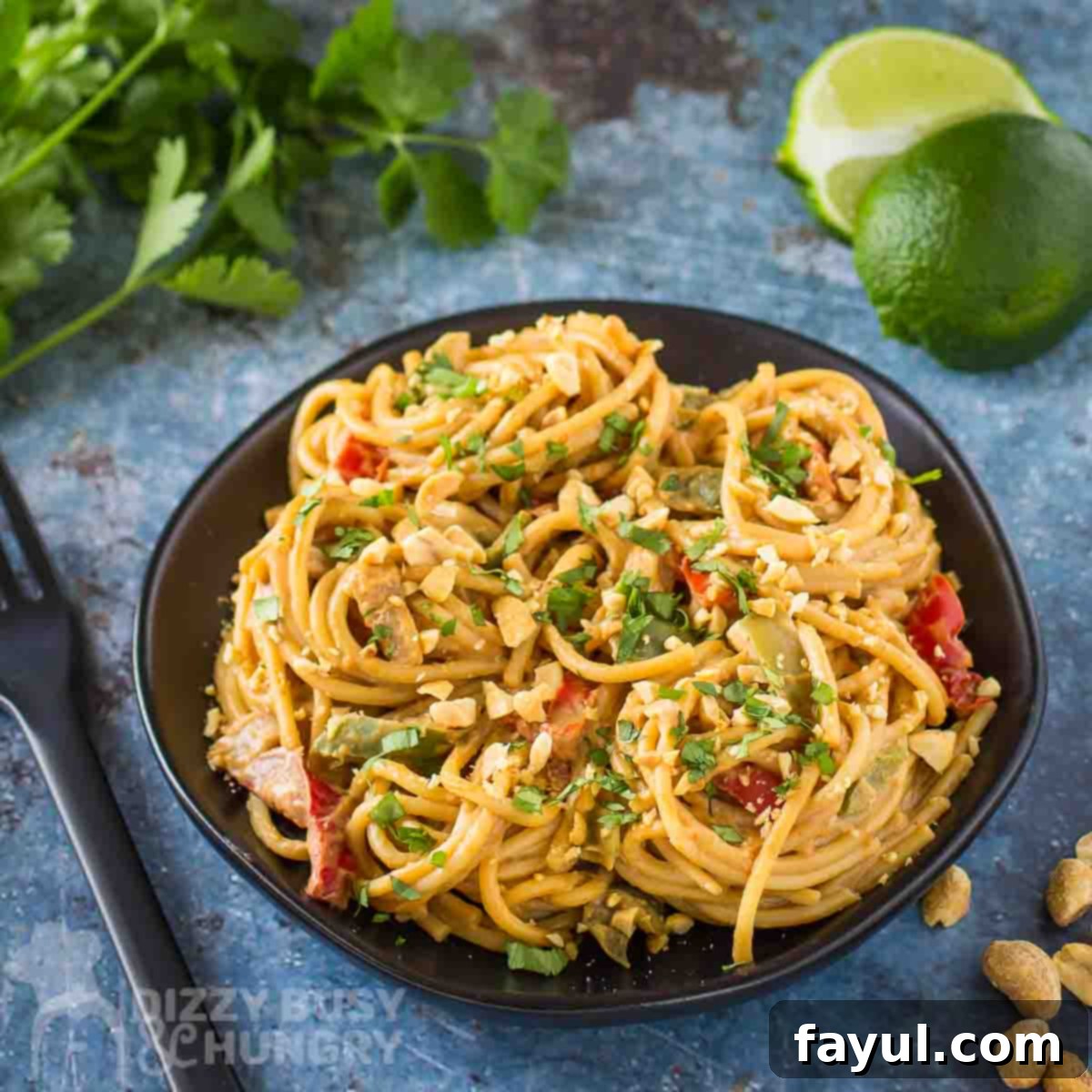 Side angled shot of vibrant Thai noodles topped with fresh cilantro and chopped peanuts on a black plate, with additional cilantro and lime wedges in the background on a blue surface, inviting a delicious meal.