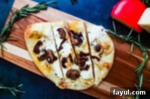 An overhead shot of the Vegetarian Flatbread Pizza recipe. The pizza is on a cutting board on a blue counter top and surrounded by fresh rosemary and 2 cheese blocks.
