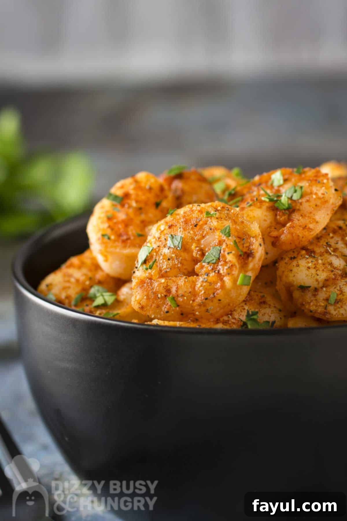 Side angled shot of sweet and spicy skillet shrimp garnished with fresh parsley in a black bowl with more herbs on the side.