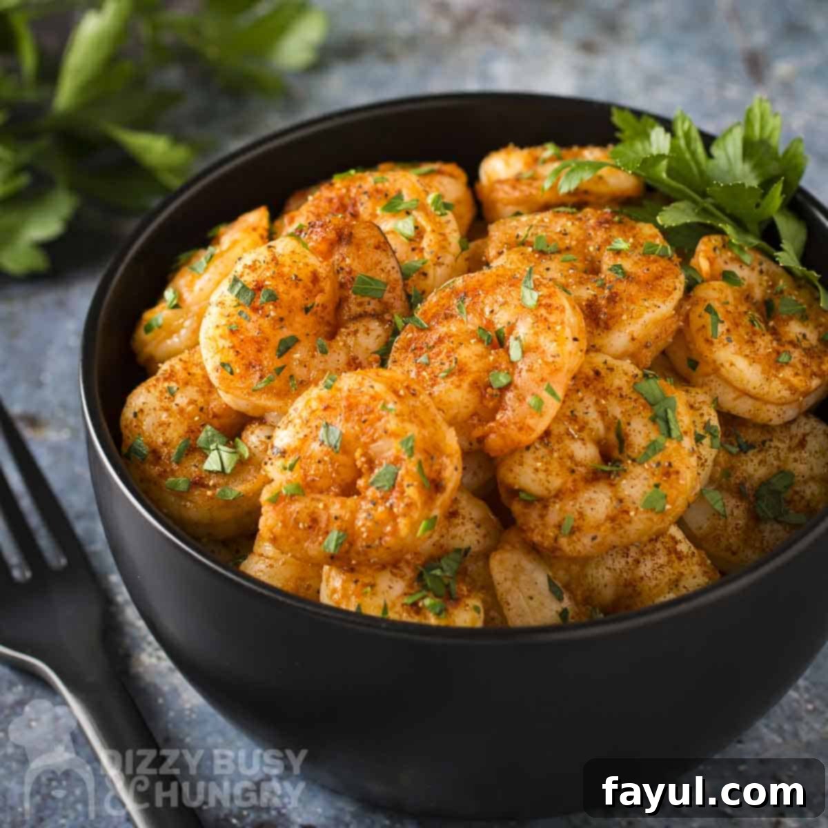 Side angled shot of sweet and spicy pan-seared shrimp garnished with fresh parsley in a black bowl with more herbs and a black fork on the side.
