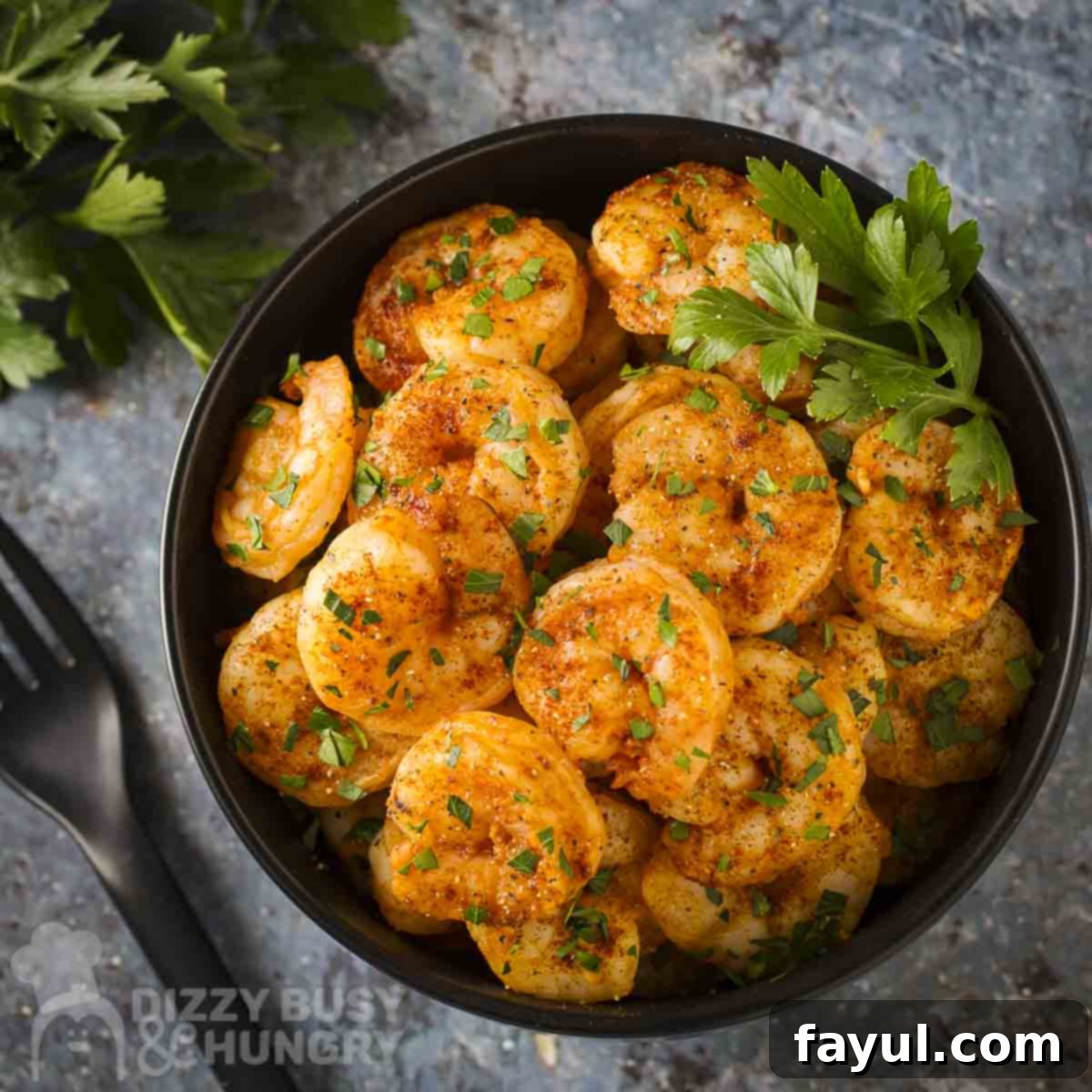 Overhead shot of sweet and spicy sriracha shrimp garnished with fresh parsley in a black bowl with more herbs and a black fork on the side.