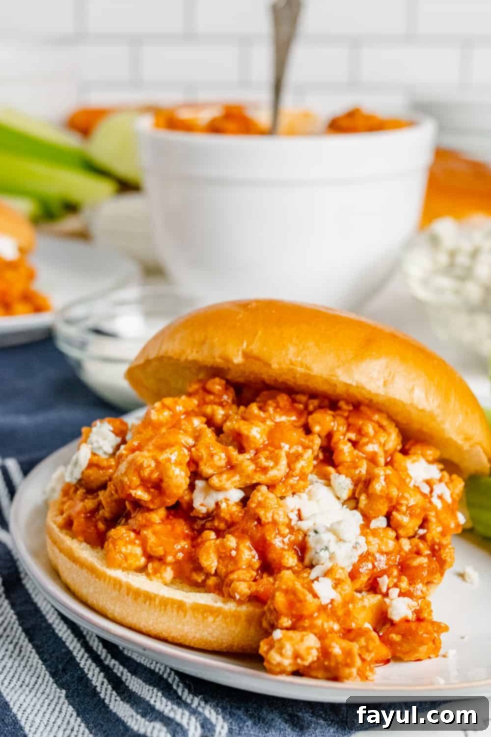 Sloppy Joe on a big white bun sitting on a white plate with a blue and white kitchen towel in the background.