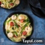 Overhead shot of shrimp pasta topped with pepper in a black bowl on a blue marble surface with more bowls and forks on the side.