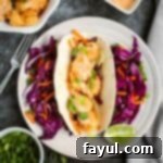 Overhead shot of one shrimp taco with shredded carrots and purple cabbage surrounding it and a lime wedge on the side of the white plate. On the black surface surrounding, there is more shrimp, sauce, and chopped green onions in white bowls as well as a fork and grey cloth.