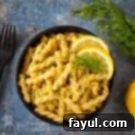 Overhead shot of pasta garnished with lemon zest, dill, and lemon slices in a black bowl on a blue surface with a black fork, dill, and a lemon on the side.