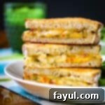 Side shot of three halves of grilled cheese sandwiches stacked on each other on a white plate with a side of greens on a blue and green cloth with a green glass in the background.