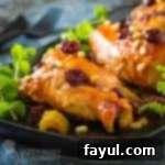 Side shot of three pieces of cranberry chicken garnished with cranberries and herbs on a black plate with cutlery in the background.
