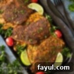 Overhead shot of blackened chicken on a black rectangle plate garnished with herbs, cherry tomatoes, and lime wedges with a black fork and knife on the side.