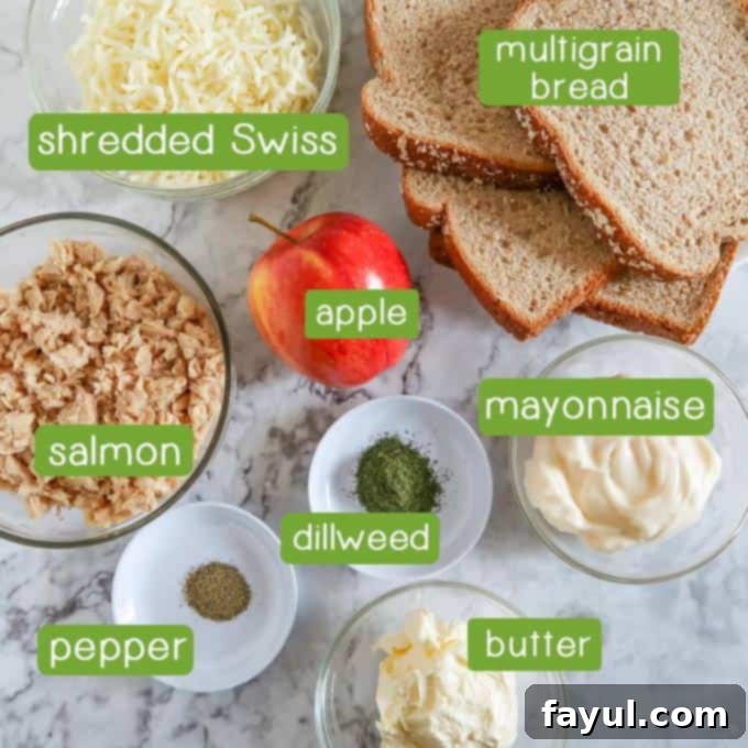 Overhead shot of fresh ingredients for gourmet grilled cheese: Shredded Swiss cheese, hearty multigrain bread, a crisp apple, canned salmon, creamy mayonnaise, fresh dill weed, black pepper, and butter.