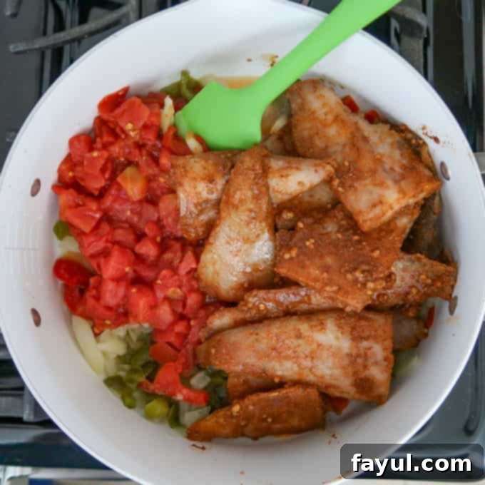 Close up shot of tomatoes and tilapia being added to onion and pepper mixture in white pan over stove.
