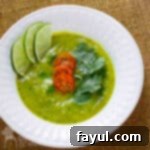 Overhead shot of zucchini curry soup garnished with tomatoes, parsley, and lime slices in a white bowl on a white plate.