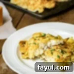 Side view of potato crusted cod on a white plate on a wooden surface with more cod on a baking sheet in the background.
