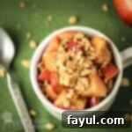 Overhead shot of a delightful apple pie snack served in a white mug, accompanied by a spoon and scattered graham cracker crumbles, all arranged on a vibrant green surface.