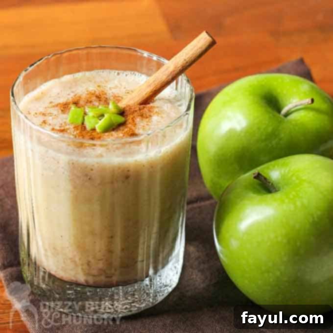 A vibrant apple smoothie with a dusting of cinnamon, fresh-cut apple pieces, and a decorative cinnamon stick on top, served in a clear glass, positioned next to two ripe granny smith apples.