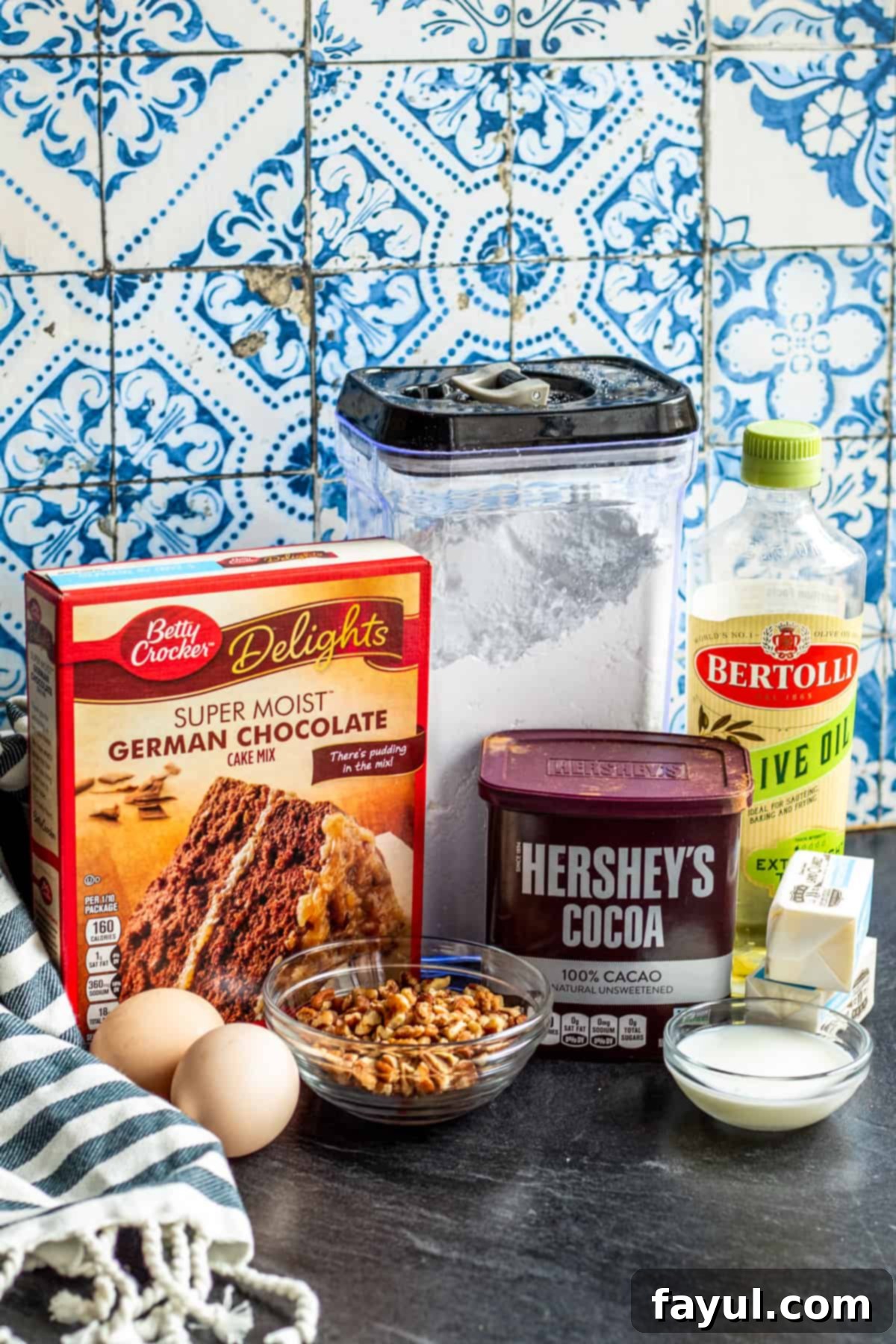 Cookie ingredients laid out on a dark counter, showing a boxed cake mix, eggs, oil, pecans, butter, milk, powdered sugar, and cocoa powder.