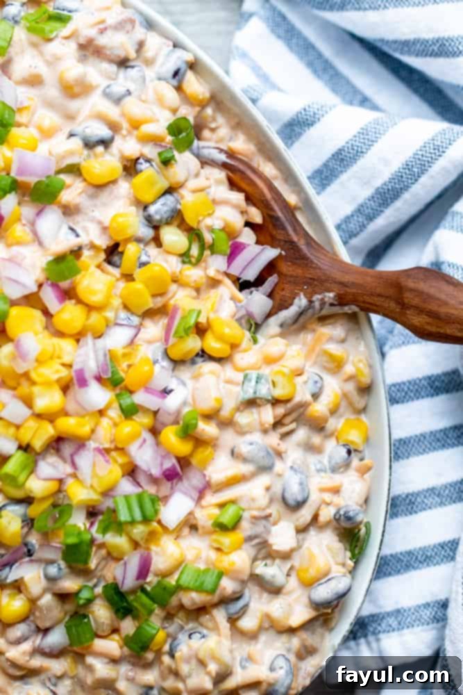 A wooden spoon scoops into a large white bowl of creamy Fiesta Corn Dip, showcasing the vibrant ingredients.