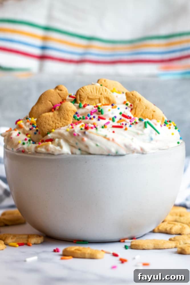 The Ultimate Funfetti Dunker Dip 2 Strait on shot of a vibrant Funfetti Cake Dip in a white bowl, adorned with colorful sprinkles, placed on a clean white countertop. Animal crackers and other dippers are artfully arranged around the bowl, inviting you to indulge in this nostalgic treat.
