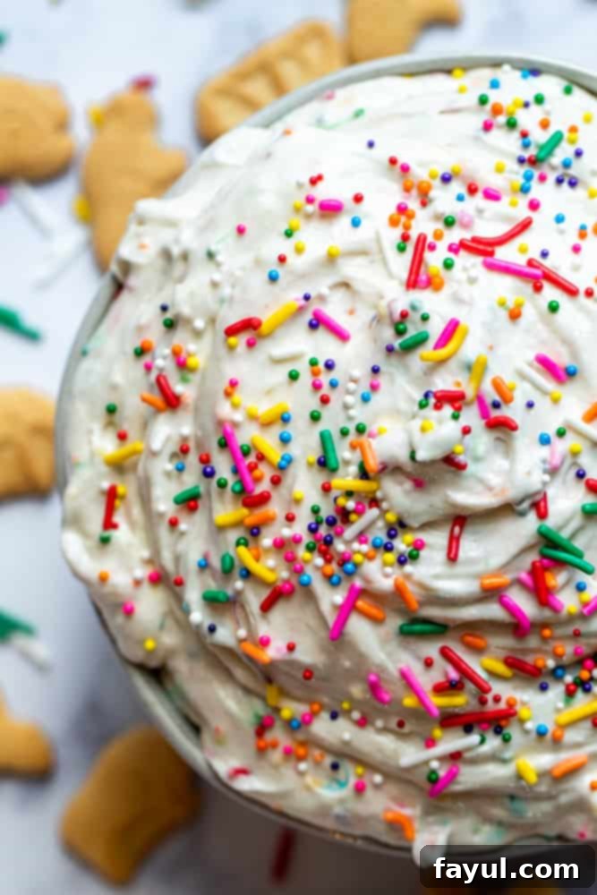 The Ultimate Funfetti Dunker Dip 5 Overhead close up shot of a fluffy Funfetti Cake Dip in a white bowl on a white counter with various dippers like animal crackers and cookies surrounding it, highlighting its vibrant colors and inviting texture.