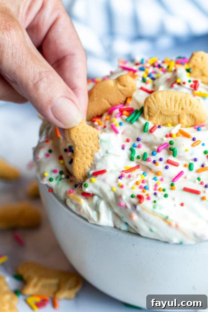 The Ultimate Funfetti Dunker Dip 6 Hand dipping animal cracker in vibrant Funfetti Cake Dip, highlighting the creamy texture and colorful sprinkles.
