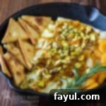 Close up shot of baked brie in a skillet with pita chips, apricots and herbs surrounding it on a wooden surface.