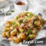Side shot of loaded tater tots on a plate with newspaper parchment with a small bowl of bacon bits in the background.